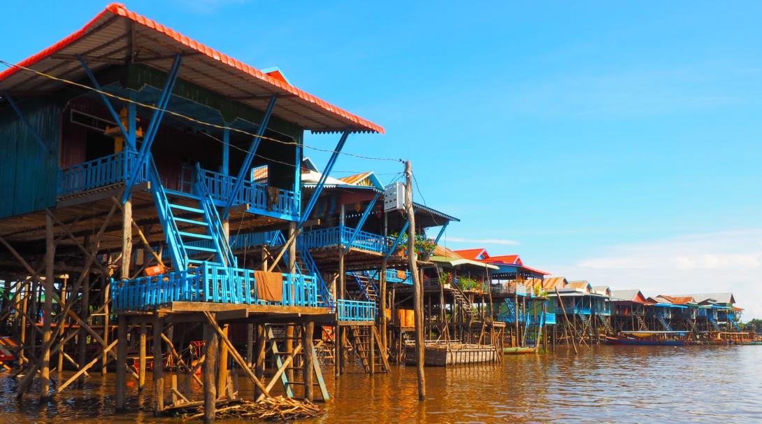 A row of blue, wooden houses stand above a still lake on long, wooden stilts