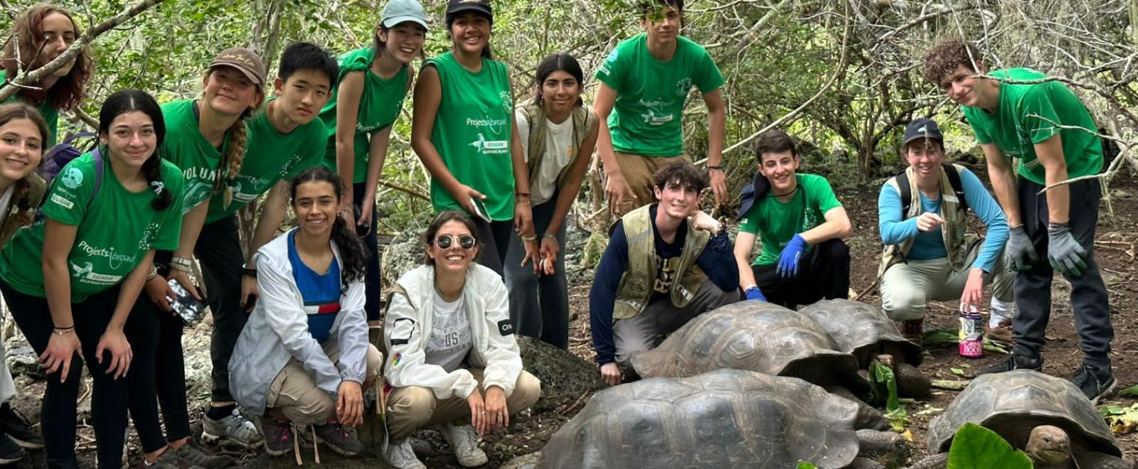 A group of young people on a trip to the Galapagos, assisting on the Projects Abroad Conservation Program. There are 15 people, some wearing green Projects Abroad t-shirts. They are crouching behind Galapagos Turtles that are eating leaves.